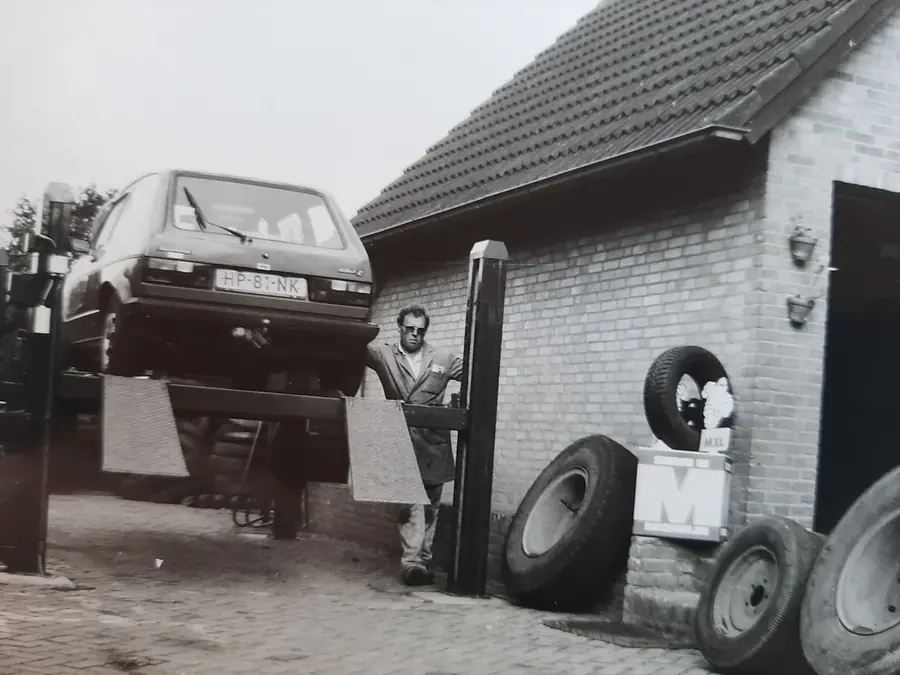 Zwart-witfoto van vroeger: een auto op de brug naast de garage, met banden tegen de muur